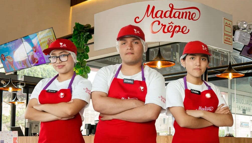 Tres integrantes del Team Madame con mandil rojo y gorra frente al mostrador de Madame Crêpe